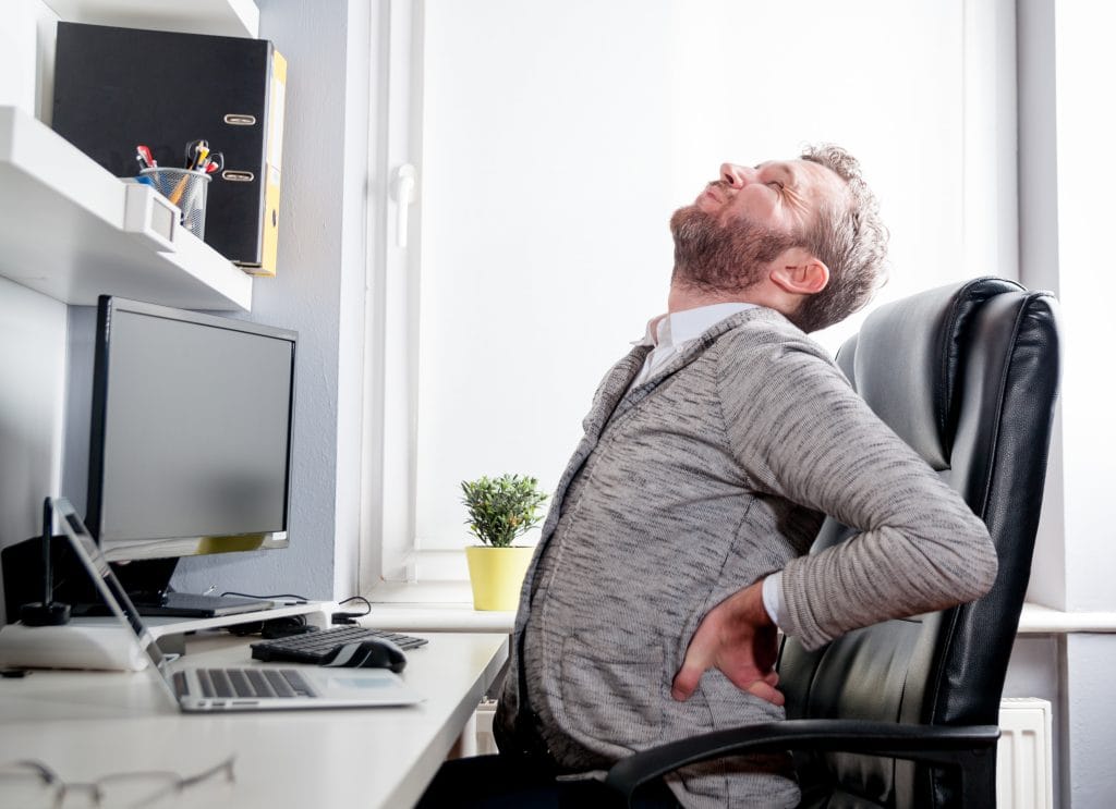 Ein Mann hält sich an einem Schreibtisch vor Schmerzen den Rücken was Rückenschmerzen am Arbeitsplatz darstellen soll